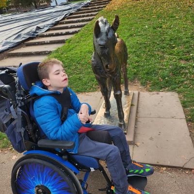 Schüler, mit blauer Jacke sitzt im Rollstuhl vor einem Bronze Pferd am Kopfende