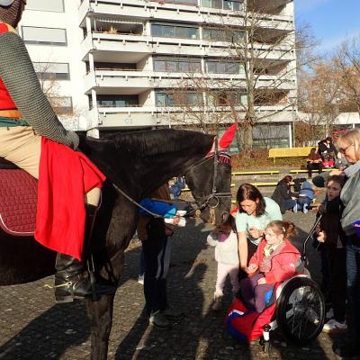 Ein Mädchen im Rollstuhl, begleitet von Erwachsenen, füttert das Pferd. Sankt Martin sitzt darauf, im Hintergrund das mehrstöckige Gebäude