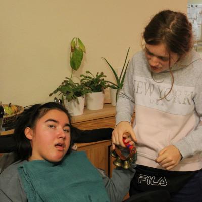 zwei junge Frauen halten gemeinsam ein Schellenband, eine davon sitzt im Rollstuhl