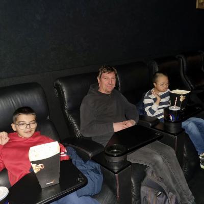 Es sitzen von links nach rechts ein Junge, ein Mann und ein weiterer Junge in gemütlichen Kinosesseln. Die Jungen haben Popcorn und ein Getränk vor sich