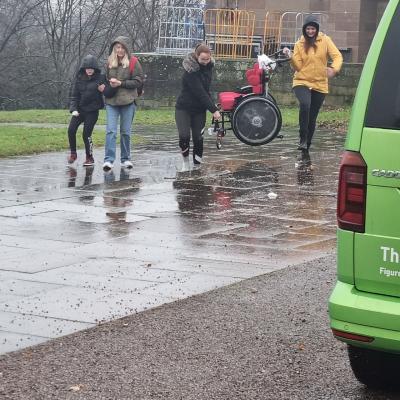 3 Erwachsene Frauen und ein Mädchen laufen durch den strömenden Regen. 2 Frauen tragen einen Rollstuhl und die andere Frau hilft dem Mädchen beim laufen. 