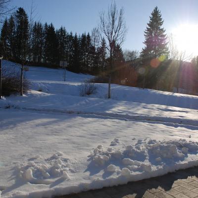 Eine schneebedeckte Landschaft wird von Abendsonne beschienen. Im Vordergrund ist ein gepflasterter Weg freigeräumt. Im Hintergrund stehen Nadelbäume.