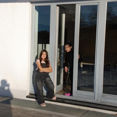 Sophia und Ghofran kommen durch die Terrassentür nach draußen. Sophia ist kurzärmlich. Die Sonne scheint beiden ins Gesicht.