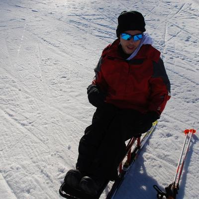 Maik sitzt im Langlaufschlitten auf dem Schnee. Neben ihm liegen auf dem Boden zwei Skistöcke.