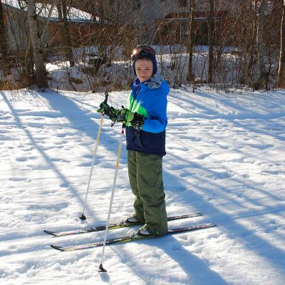 Luca läuft in der Loipe und lächelt in die Kamera. Hinter ihm sieht man Bäume, die Schatten auf den Schnee werfen. Dahinter sind Häuser.