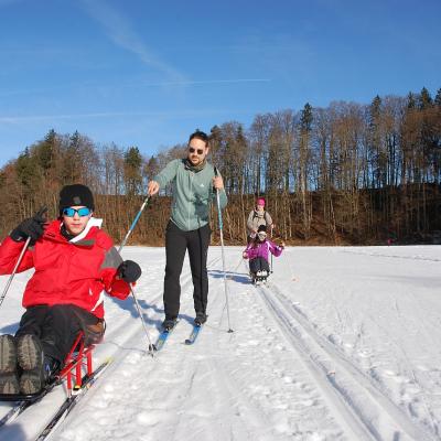 Man sieht zwei Loipen frontal. Links kommt Maik im Schlitten unterstützt von Herr Kling. Rechts etwas weiter hinten kommt Marija im Schlitten unterstützt von Frau Gauch. Im Hintergrund sieht man Bäume.