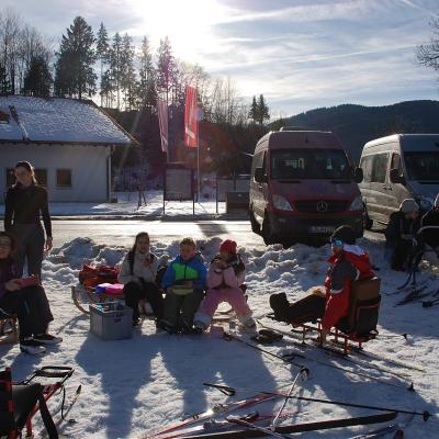 Die Schüler:innen sitzen auf verschiedenen Schlitten und essen ihr Vesper. Frau Gauch und Lena stehen daneben. Im Hintergrund sieht man die geparkten Busse, ein Haus, zwei Flaggen. 