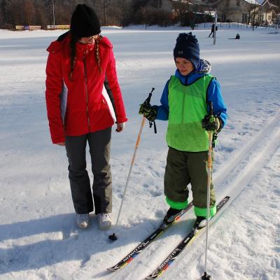 Luca fährt in der Loipe. Neben ihm läuft Lena und schaut ihn an. Die Sonne wirft ihre Schatten neben sie in den Schnee.