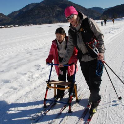 Ghofran läuft am Skilator und Frau Gauch hält den Skilator vorne. Dahinter sieht man bewaldete Berge und blauen Himmel.
