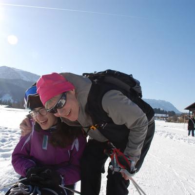 Marija sitzt im Skischlitten. Frau Gauch beugt sich herunter und legt ihr den Arm um die Schulter. Beide lachen. Im Hintergrund Berge und blauer Himmel.