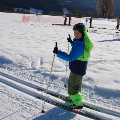 Luca läuft in der Loipe und lächelt in die Kamera. Am Kopf hängt ein hellgrünes Leibchen.