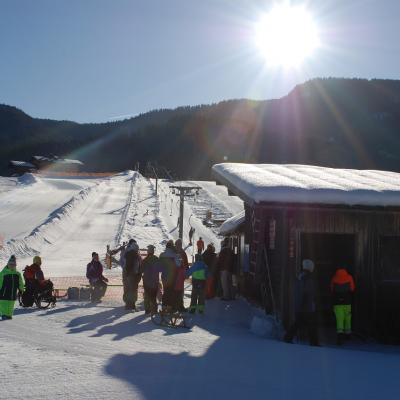 An der Hütte des Lifts stehen alle Schüler: innen teilweise in der Schlange. Dahinter sieht man bewaldete Berge, blauen Himmel und Sonne.