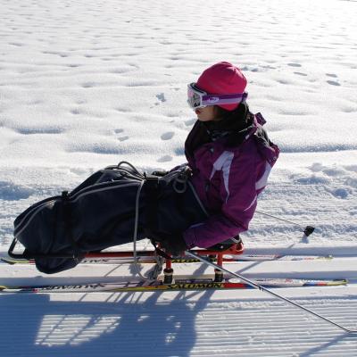 Marija sitzt im Schischlitten auf der Loipe und stochert mit den Stöcken rechts und links. Dahinter sieht man schneebedeckten Boden.