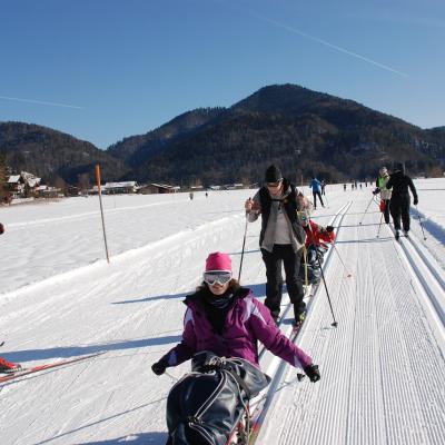In einer Loipe kommen Marija, Frau Gauch und Maik angefahren. Auf der anderen Spur  und daneben laufen andere Menschen. Dahinter Berge.