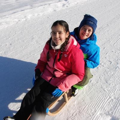 Ghofran sitzt vor Luca auf einem Holzschlitten auf dem Schneeweg. Beide lächeln in die Kamera.