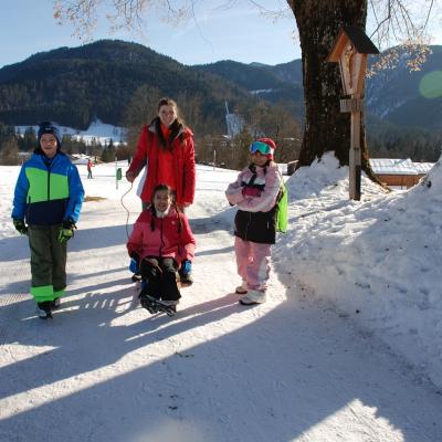 Auf einem schneebedeckten Weg kommen Luca, Lene, Sophia und Ghofran der Kamera entgegen. Ghofran sitzt auf einem Holzschlitten. Alle lächeln in die Kamera.