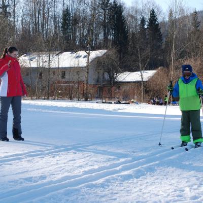 Luca läuft in der Loipe. Lena steht daneben und feuert ihn mit einer Rätsche an. Dahinter Häuser, Bäume, einen Berg und Himmel sowie andere Kinder.