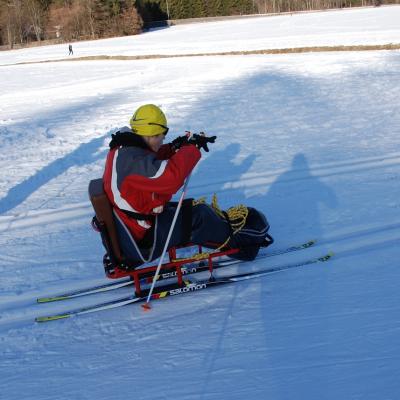 Maik stochert im Skischlitten in der Loipe. Dahinter sieht man Schnee und Bäume.