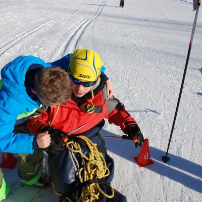 Maik steht mit dem Skischlitten in der Loipe auf der Ziellinie zwischen zwei Pelonen., Luca beugt sich zu ihm herunter und umarmt ihn gratulierend.