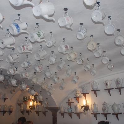 An der Decke eines gewölbten Raums hängen viele verschiedene Kannen. Auch auf Regalbrettern an den Wänden stehen Kannen.