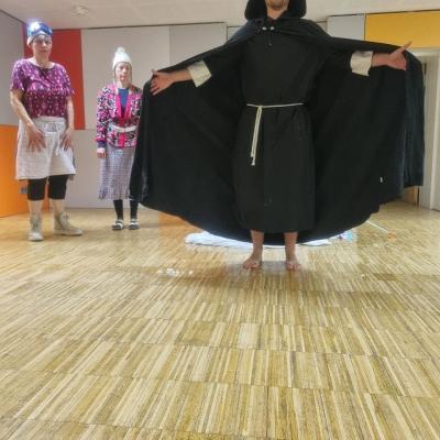 Der Hohepriester hat die Hände ausgebreitet. Er trägt Kutte und Umhang. Leni und Berta stehen hinter ihm. Sie tragen bunte Kleidung und eine Skibrille.