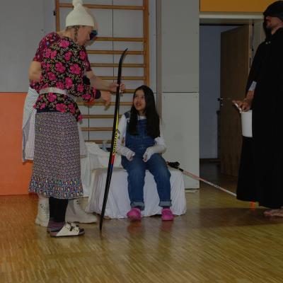 Leni, Berta und der Hohepriester stehen um Ghofran, die auf dem weißen Thron sitzt. Leni hält einen Langlaufski. Der Hohepriester hält einen Eimer.