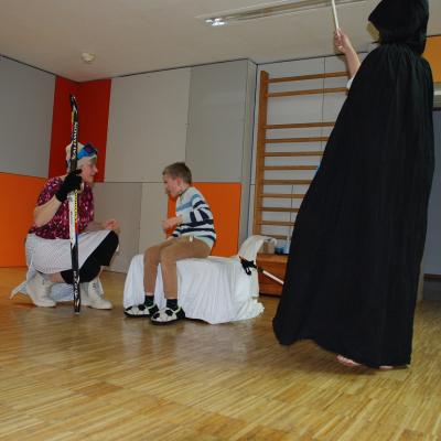 Leni, Berta und der Hohepriester stehen um Luca der auf dem weißen Thron sitzt. Der Priester spritzt ihn mit Wasser nass, Leni hält die Urkunde, Berta den Ski.