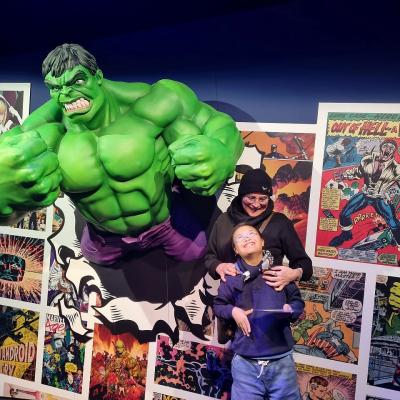 Eine Frau und ein Junge stehen vor einer Wand und lachen. Neben ihnen kommt ein große grüne Figur Namens Hulk aus der Wand. 