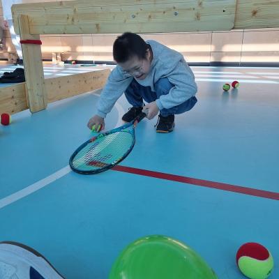 Ein Junge ist in einer Sporthalle in der Hocke. Er hält einen Tennisschläger und greift mit der anderen Hand nach einem Tennisball