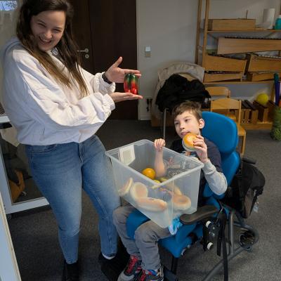 Ein Junge im Rollstuhl spiel mit einer Kiste voller Rasseln und eine Frau lacht lustig in die Kamera