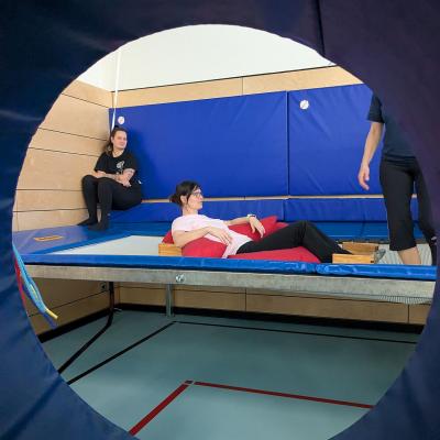 Zwei Frauen sitzen auf dem Trampolin, eine am Rand, eine andere auf dem Sprungtuch in einem Kasten