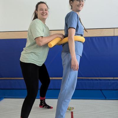 Zwei Frauen hüpfen auf einem Trampolin, eine Person wird durch einen Ring unterstütz 