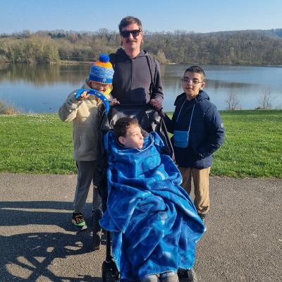 Ein Mann steht mit drei Jungen vor einem See. Ein Junge sitzt im Buggy. Die Sonne scheint. 