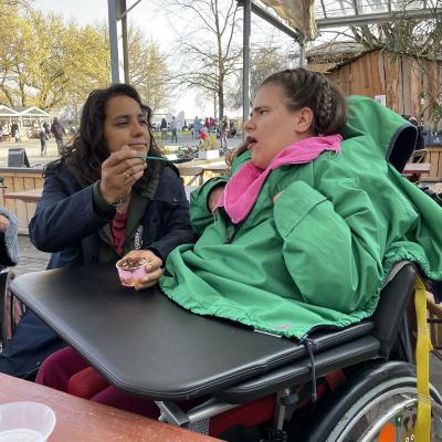 Eine Frau reicht einer jungen Frau im Rollstuhl ein Schokoladeneis