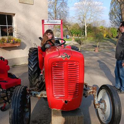 Schülerin sitzt lachend auf einem roten Traktor und hält das schwarze Lenkrad fest. Am hinteren Teil vom Traktor steht die Aufschrift 