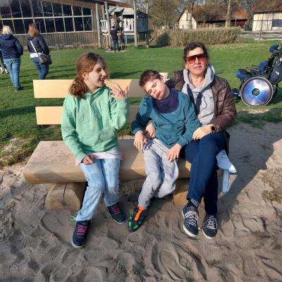 eine Betreuerin und zwei Schüler sitzen auf einer hellen Holzbank, die Holzbank steht im Sandkasten, im Hintergrund rechts steht ein leerer Rollstuhl