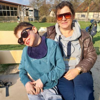 Betreuerin und Schüler sitzen auf der hellen Holzbank  mit Sonnebrille, beide lächeln in die Kamera