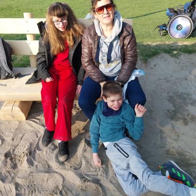 aneben eine Schülerin mit zwei Zöpfen, vor der Betreuerin sitzt ein Schüler im Sand angelehnt an den Beinen, er lacht