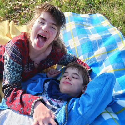 Schüler mit blauer Jacke liegt entspannt auf dem Rücken, Schülerin rechts mit rotem Oberteil schaut in die Kamara und streckt die Zunge raus 