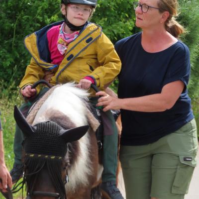 Ein 9jähriges Mädchen mit Helm frontal abgebildet auf einem dunkelbraun-weiß gefleckten Pony, daneben eine Frau als Begleitung. Im Hintergrund grün belaubte Bäume.