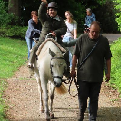 Ein neunjähriger Junge mit Helm frontal aufgenommen auf einer weißen Stute im Wald, begleitet von einem Mann und einer Frau rechts neben ihm. Im Hintergrund 4 weitere Erwachsene.