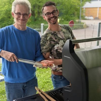 Zwei Männer stehen am Grill und grillen, lachen in die Kamera
