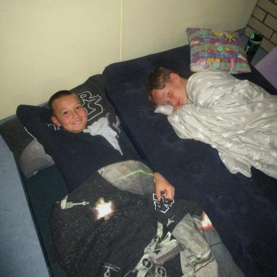 2 nebeneinander liegende Jungen auf Luftdmatratzen im Klassenzimmer, in Decken gehüllt.