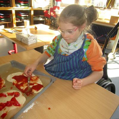 Ein Mädchen im Rollstuhl verteilt im Klassenzimmer mit einem Löffel Tomatensoße auf Pizzateig