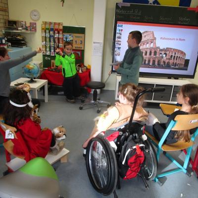 2 Schüler spielen einen Schwertkampf der Gladiatoren, davor als Raubkatzen verkleidete Kinder. Im Hintergrund ein Legamaster mit einer Abbildung des Kolosseums.
