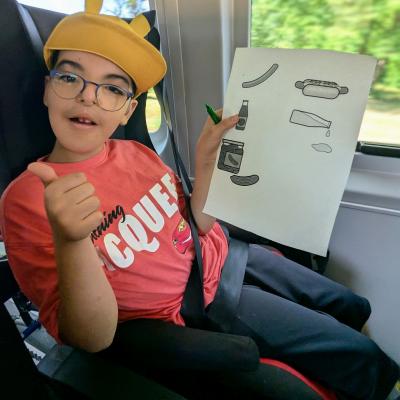 ein Junge sitzt im Auto, zeigt seine Einkaufsliste und lacht in die Kamera