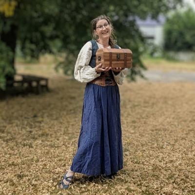 Eine Frau als Hofdame verkleidet lacht in die Kamera und hält eine Kiste in der Hand 
