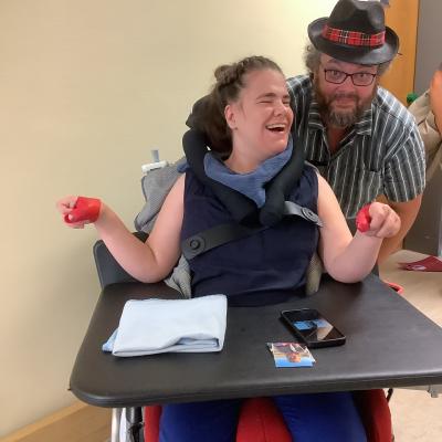 Eine junge Frau im Rollstuhl steht neben einem Mann, beide lachen. Der Mann trägt einen Hut.