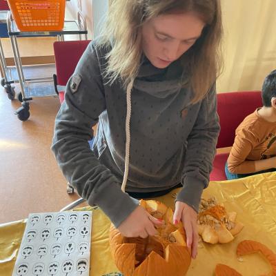 Ein Mädchen steht vor dem Tisch. Vor ihr ist ein Kürbis. Der Kübis hat oben ein Loch. Die Kanten sind zackig. Sie holt das Fruchtfleisch und die Kerne aus dem Kürbis